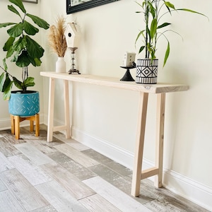 May include: A light wood console table with a long, narrow top and two sets of angled legs. The table is decorated with a white birdhouse, a vase with dried grasses, a white vase, and a black and white patterned planter with a green plant.