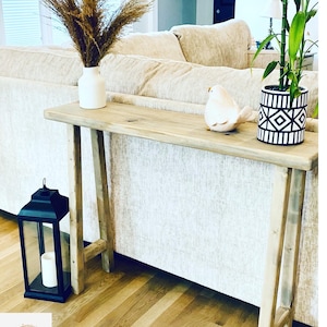 May include: A wooden console table with a light wood finish. The table has a simple design with two legs on each side. The table is decorated with a vase of dried pampas grass, a white ceramic bird figurine, and a potted plant with black and white geometric patterned pot.
