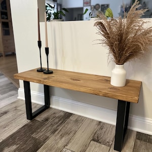 May include: A wooden bench with a natural finish, supported by black metal legs. The bench is adorned with two black candle holders with candles and a white vase filled with dried pampas grass. The bench is approximately 48 inches long.