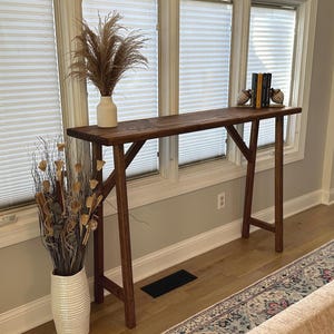 May include: A dark brown wooden console table with a long, narrow top and a simple A-frame base. The table is positioned in front of a window with white blinds. A white vase with dried pampas grass sits on the table, and a set of books is propped against a pair of decorative bird bookends.