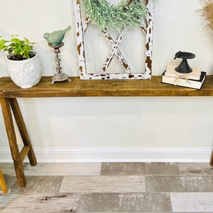 May include: A rustic wooden console table with a brown stain finish. The table has a simple A-frame design and is decorated with a white owl planter, a bird figurine, a wreath, and a stack of books.