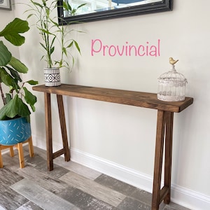 Puede incluir: Una mesa consola de madera marr&oacute;n oscuro con un acabado r&uacute;stico. La mesa tiene una tapa rectangular y dos patas en forma de A. La mesa est&aacute; decorada con una planta en maceta y una jaula para p&aacute;jaros con una peque&ntilde;a figura de p&aacute;jaro. El texto "Provincial" est&aacute; escrito en rosa en la pared detr&aacute;s de la mesa.