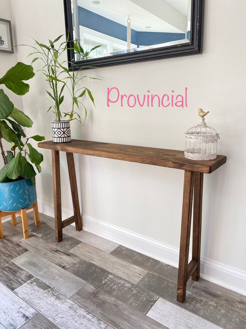 Puede incluir: Una mesa consola de madera marr&oacute;n oscuro con un acabado r&uacute;stico. La mesa tiene una tapa rectangular y dos patas en forma de A. La mesa est&aacute; decorada con una planta en maceta y una jaula para p&aacute;jaros con una peque&ntilde;a figura de p&aacute;jaro. El texto "Provincial" est&aacute; escrito en rosa en la pared detr&aacute;s de la mesa.