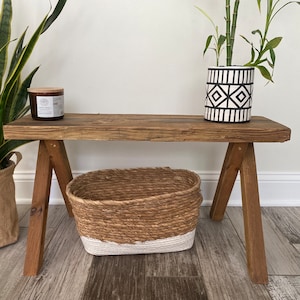 May include: A rustic wooden bench with a natural wood finish. The bench has two A-frame legs and a wide, flat top. A woven basket and a candle are on the bench. A potted plant with a black and white geometric pattern is on the bench.