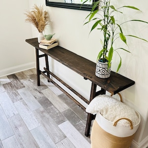 May include: A dark brown wooden console table with a white vase and dried flowers, a small bird figurine, and a stack of books. A potted plant with green leaves and a black and white geometric pot sits on the table. A white and beige woven basket with a white blanket sits on the floor next to the table.