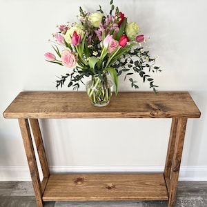 May include: A wooden console table with a single shelf. The table is stained a dark brown color and has a rustic look. A vase of pink, white, and green flowers sits on the top shelf.