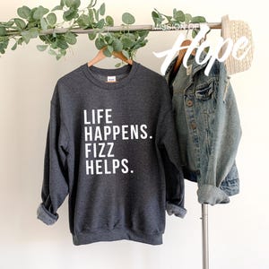 May include: A dark gray sweatshirt with the text "Life Happens. Fizz Helps." printed in white on the front.
