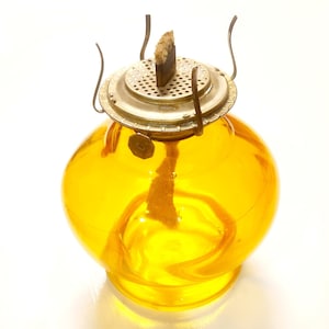 Puede incluir: Una lámpara de aceite vintage con un cuerpo de vidrio amarillo redondo y una parte superior de latón. La lámpara presenta una mecha y una pequeña etiqueta de metal. El vidrio tiene una ligera transparencia. La lámpara está diseñada para colocarse sobre una superficie plana.