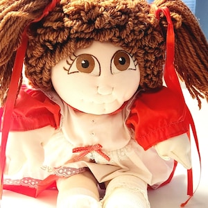May include: A handmade doll with brown yarn hair styled in pigtails, tied with red ribbons. The doll has a painted face with large brown eyes and is wearing a red and white dress with a red bow. The doll's legs are white.