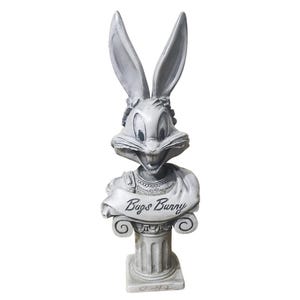 Könnte beinhalten: Graue Bugs Bunny Büste. Die Statue zeigt Bugs Bunnys Kopf und Oberkörper mit einem Banner mit der Aufschrift "Bugs Bunny". Die Statue ist auf einem Säulensockel montiert.