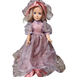 May include: A vintage doll with blonde hair and blue eyes, wearing a pink dress with floral patterns and a matching bonnet. The dress features sheer sleeves and a long, flowing skirt. A pink bow adorns the waist.