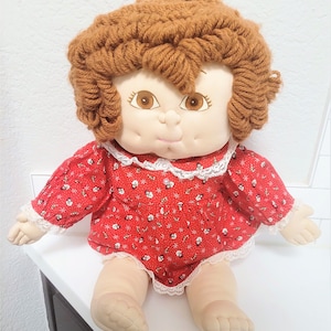 May include: A vintage rag doll with brown yarn hair and embroidered facial features. The doll wears a red patterned dress with white lace trim. The doll is sitting on a white surface.