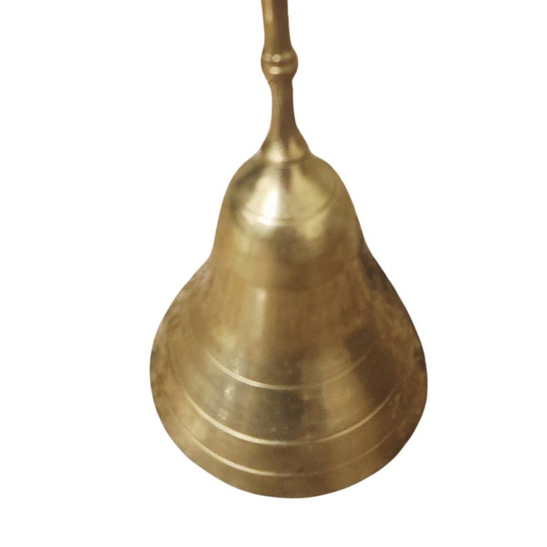 Puede incluir: Una campana dorada brillante con un mango largo. La campana tiene forma redondeada con tres ranuras horizontales cerca de la base. El mango es delgado y se curva ligeramente en la parte superior. La campana es de metal y tiene un acabado pulido.