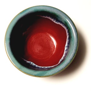 Può includere: Una ciotola in ceramica fatta a mano con interno rosso lucido e bordo turchese. Il bordo ha un accento blu scuro e bianco. La ciotola è rotonda e poggia su una superficie bianca. Questa ciotola decorativa è ideale per servire cibo.