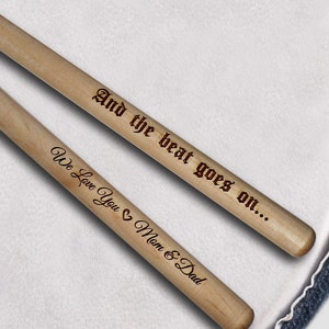 Peut inclure: Une paire de baguettes de batterie en bois avec le texte "And the beat goes on... We Love You Mom & Dad" grav&eacute; dessus.