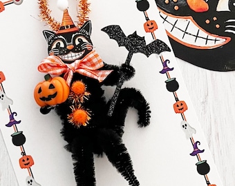 May include: A black cat with a pumpkin and a witch's hat made of pipe cleaners. The cat is holding a small orange pumpkin in its paws. The cat is surrounded by a border of orange pumpkins and ghosts. The text "APRIL & KIWI" is at the bottom of the image.