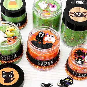 May include: A selection of Halloween-themed slime jars with black lids and decorative labels. The jars contain green and orange slime, decorated with Halloween charms such as ghosts, spiders, and pumpkins. Each jar has a name label.