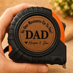 Puede incluir: Un flex&oacute;metro naranja y negro con el texto "No One Measures Up To You! DAD" y los nombres "Harper & Jax" grabados en una placa circular marr&oacute;n. El flex&oacute;metro est&aacute; en una mano.