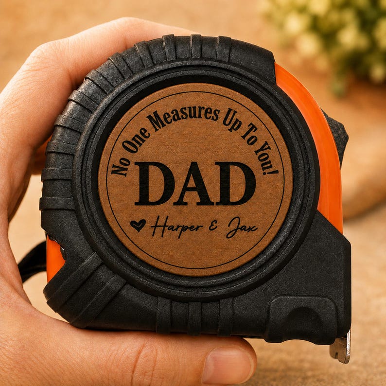 Puede incluir: Un flex&oacute;metro naranja y negro con el texto "No One Measures Up To You! DAD" y los nombres "Harper & Jax" grabados en una placa circular marr&oacute;n. El flex&oacute;metro est&aacute; en una mano.