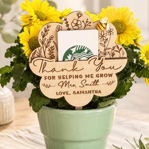 May include: A wooden flower-shaped gift tag with the words "Thank You" and "For helping me grow Mrs. Smith Love, Samantha" engraved on it. The tag is placed in a green pot with yellow flowers and a Starbucks gift card.