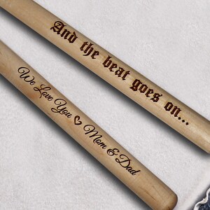 Peut inclure: Deux baguettes de batterie en bois avec le texte "And the beat goes on..." sur une baguette et "We love you Mom & Dad" sur l'autre baguette.