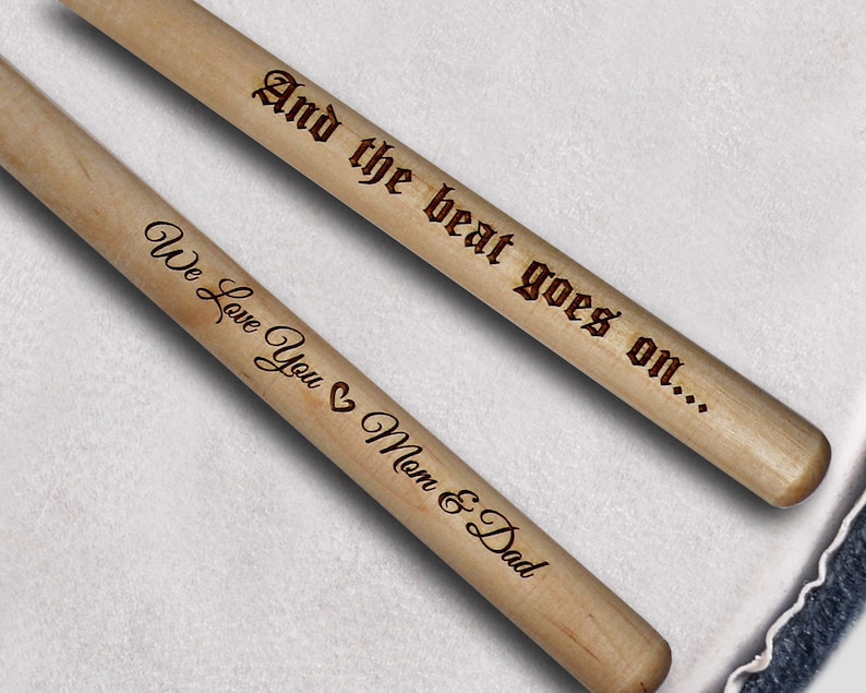 Peut inclure: Deux baguettes de batterie en bois avec le texte "And the beat goes on..." sur une baguette et "We love you Mom & Dad" sur l'autre baguette.