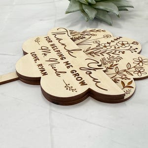 May include: A wooden, four-leaf clover-shaped sign with the words "Thank You For Helping Me Grow Ms. Nicole Love, Ryan" engraved in a cursive font. Floral designs are etched on the clover leaves. The sign is light brown.