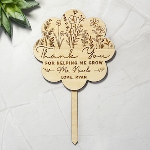 May include: A wooden flower-shaped garden stake with engraved floral designs and the words "Thank You For Helping Me Grow Ms. Nicole Love, Ryan". The stake is made of light-colored wood.