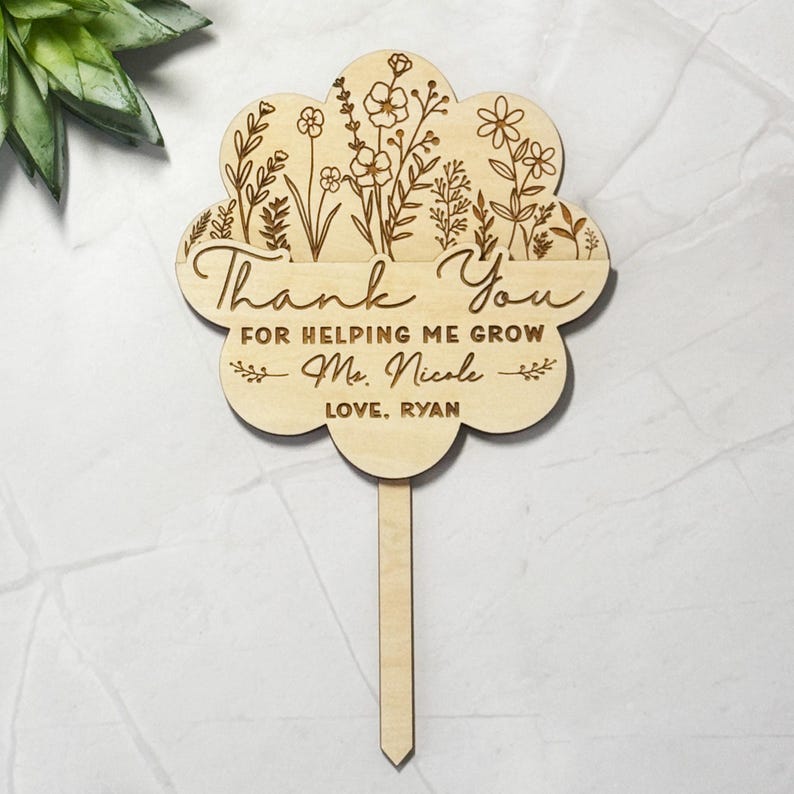 May include: A wooden flower-shaped garden stake with engraved floral designs and the words "Thank You For Helping Me Grow Ms. Nicole Love, Ryan". The stake is made of light-colored wood.
