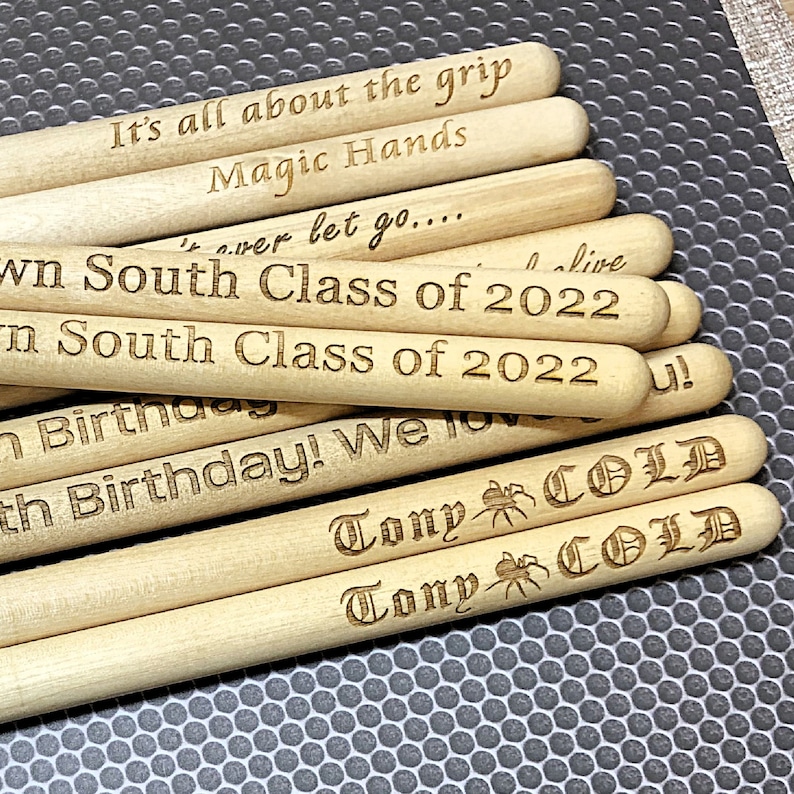 Peut inclure: Une pile de b&acirc;tonnets en bois avec diff&eacute;rentes phrases grav&eacute;es dessus, notamment "It's all about the grip", "Magic Hands", "South Class of 2022", "Birthday! We love you!" et "Tony COLD".