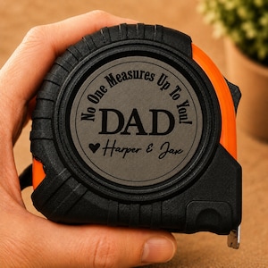 Puede incluir: Cinta m&eacute;trica negra y naranja con el texto "No One Measures Up To You! DAD Harper & Jax" grabado en el centro. La cinta m&eacute;trica se sostiene en una mano y es un regalo para un padre.