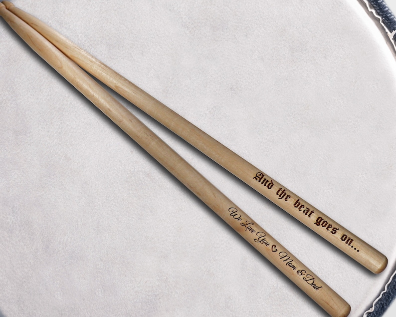 Peut inclure: Une paire de baguettes de batterie en bois avec le texte "And the beat goes on... We Love You Mom & Dad" grav&eacute; dessus.