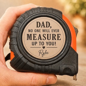 Puede incluir: Un flex&oacute;metro negro y naranja con el texto "DAD, NO ONE WILL EVER MEASURE UP TO YOU!" y el nombre "Ryfee" impreso en un c&iacute;rculo beige. El flex&oacute;metro est&aacute; en una mano.