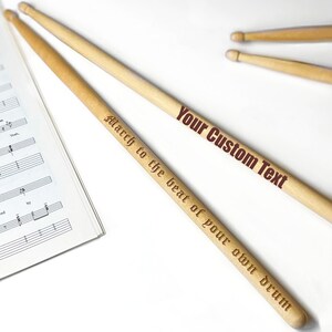 Peut inclure: Deux baguettes de batterie en bois avec le texte "Your Custom Text" et "March to the beat of your own drum" grav&eacute; dessus.