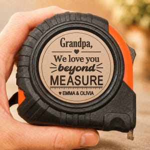 Puede incluir: Cinta m&eacute;trica negra y naranja con el texto "Grandpa, We love you beyond MEASURE" y los nombres "Emma & Olivia". La cinta m&eacute;trica se sostiene en una mano.