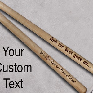 Peut inclure: Deux baguettes de batterie en bois avec des gravures personnalis&eacute;es. La baguette du haut porte l'inscription "And the beat goes on..." et la baguette du bas porte l'inscription "We Love You Mom & Dad".