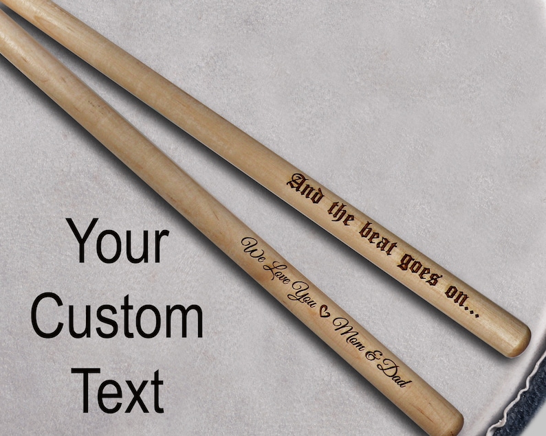 Peut inclure: Deux baguettes de batterie en bois avec des gravures personnalis&eacute;es. La baguette du haut porte l'inscription "And the beat goes on..." et la baguette du bas porte l'inscription "We Love You Mom & Dad".