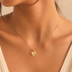 May include: A gold-colored necklace with a delicate chain and a heart-shaped pendant. The pendant features the letter "B" in a script font. The necklace is worn around the neck.