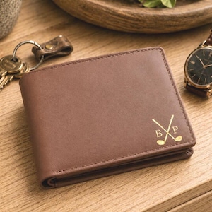 May include: Brown leather wallet with gold golf club graphic and initials. The bi-fold wallet is on a wooden surface with a watch and keys in the background. The wallet has a classic design.
