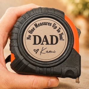 Puede incluir: Cinta m&eacute;trica negra y naranja con el texto "No One Measures Up To You! DAD Kami" en un dise&ntilde;o circular. La cinta m&eacute;trica est&aacute; sujeta en una mano.