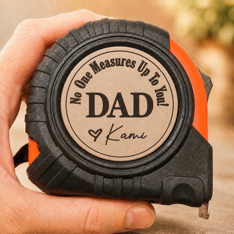 Puede incluir: Cinta m&eacute;trica negra y naranja con el texto "No One Measures Up To You! DAD Kami" en un dise&ntilde;o circular. La cinta m&eacute;trica est&aacute; sujeta en una mano.