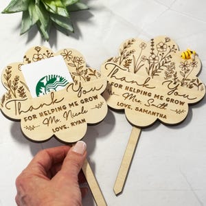 May include: Two wooden flower-shaped gift card holders with engraved floral designs and the words "Thank You." One holds a gift card. The text reads "For helping me grow" and includes names. A small bee is on one.
