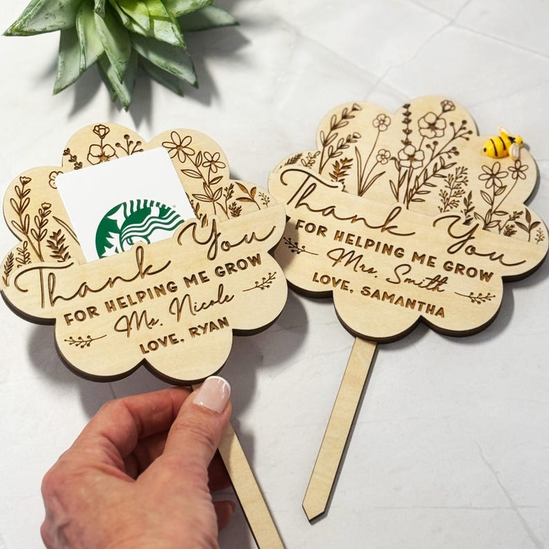 May include: Two wooden flower-shaped gift card holders with engraved floral designs and the words "Thank You." One holds a gift card. The text reads "For helping me grow" and includes names. A small bee is on one.