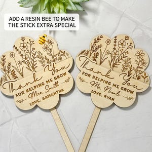 May include: Two wooden flower-shaped teacher appreciation gifts with the words "Thank You" and "For Helping Me Grow" engraved. Each has a name and a small wooden stick. One has a resin bee attached. The text "Add a resin bee to make the stick extra special" is at the top.