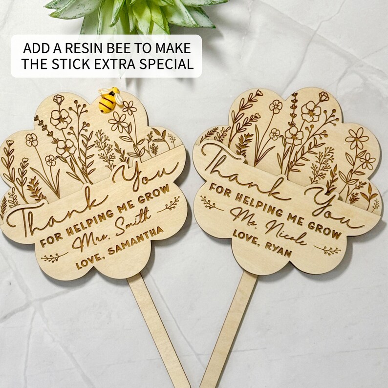 May include: Two wooden flower-shaped teacher appreciation gifts with the words "Thank You" and "For Helping Me Grow" engraved. Each has a name and a small wooden stick. One has a resin bee attached. The text "Add a resin bee to make the stick extra special" is at the top.