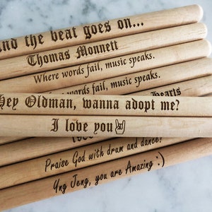 Peut inclure: Un ensemble de baguettes de batterie en bois avec des messages personnalis&eacute;s grav&eacute;s dessus. Les messages incluent "And the beat goes on...", "Thomas Monnett", "Where words fail, music speaks.", "Hey Oldman, wanna adopt me?", "I love you", "Praise God with drum and dance!" et "Ung Jeng, you are Amazing :)".