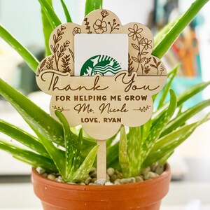 May include: A wooden flower-shaped gift card holder with the words "Thank You" and a personalized message. The holder is placed in a potted aloe vera plant, with a Starbucks gift card visible. The design includes floral engravings.