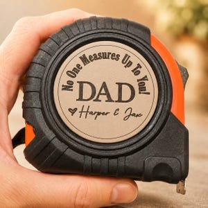 Puede incluir: Cinta m&eacute;trica negra y naranja con el texto "No One Measures Up To You!" y "DAD" con un s&iacute;mbolo de coraz&oacute;n y los nombres "Harper & Jax". La cinta m&eacute;trica est&aacute; en una mano.