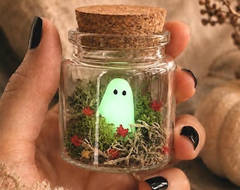 May include: A small glass jar with a cork stopper, containing a glowing green ghost figure. The jar is filled with moss and small red and orange leaves. The jar is held in a hand with black nail polish.