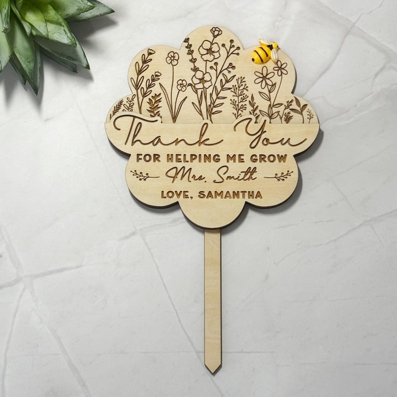 May include: A wooden flower-shaped garden stake with engraved floral designs and the words "Thank You For Helping Me Grow Mrs. Smith Love, Samantha." A small, yellow and black bee is attached to the top. The stake is light brown.